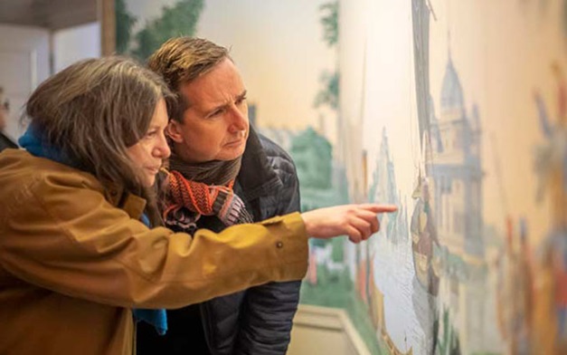 A man and a woman, dressed for cold weather, look closely at a painting. The woman is pointing at a specific detail.