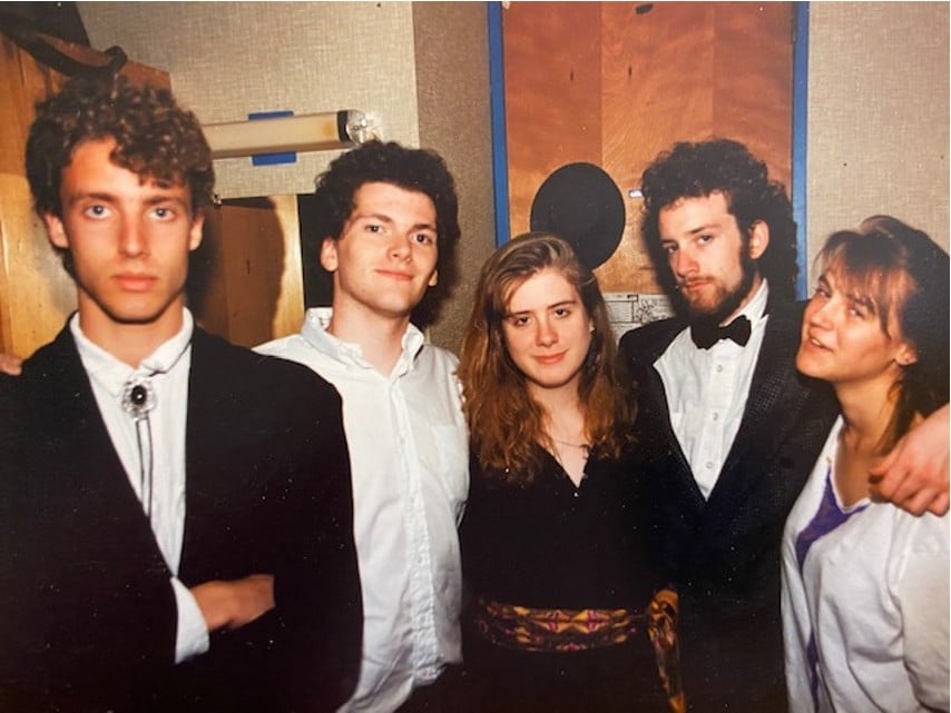 photo of 5 college students heading out to some event, circa 1990 or 1991: there's a bearded guy in a tux 2nd from right, with his arm around two lovely women. On the left, there's a guy making a dramatic model face, wearing a wrinkled dress shirt with a goddamned bolo tie. He's got a heck of a jawline, admittedly.