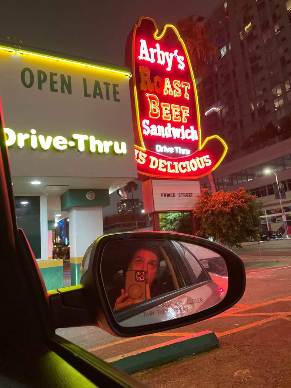 An old Arby’s neon sign glowing in the dark and a person reflected in a side view mirror of a car taking a picture of the sign
