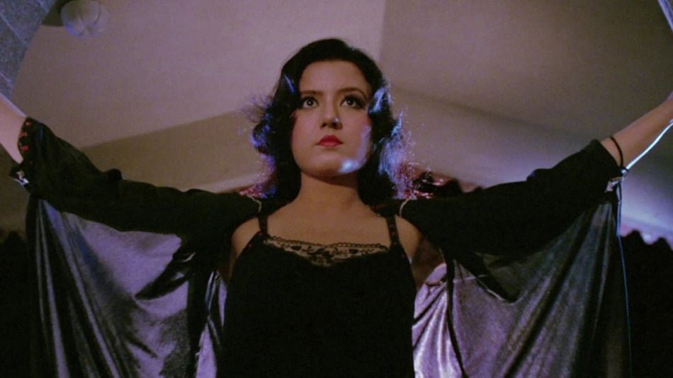 Jasmin as Veerana, standing with her arms raised, wearing a black lacy spaghetti strap number with a black cloak to cover her bare shoulders. She doesn't look too pleased.