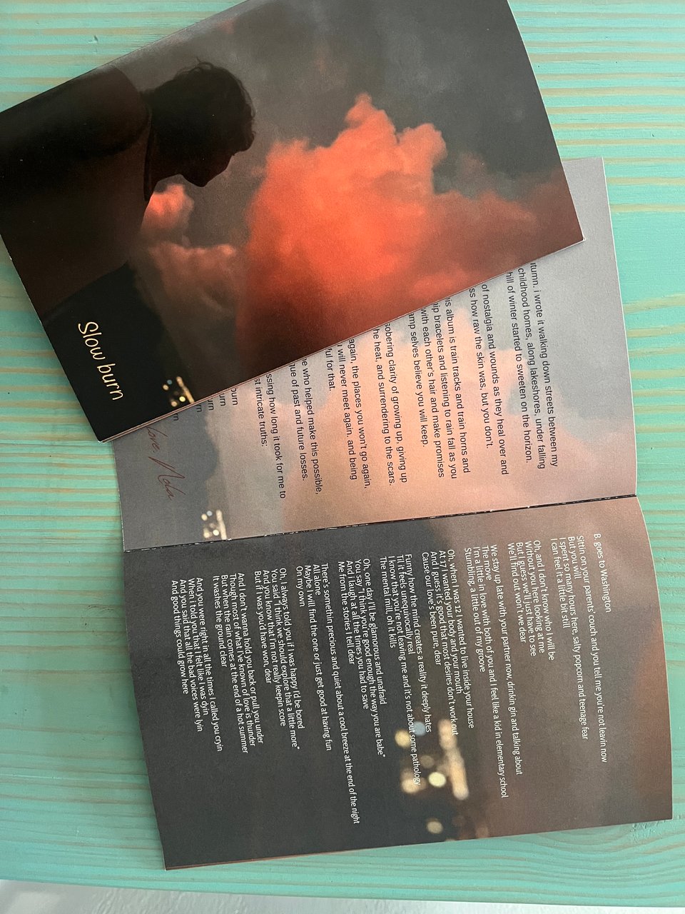two Slow burn lyric books against turquoise wood. one is closed and shows the album art of Nola in shadow looking up at vivid rose gold clouds in blue-grey sky, with Slow burn written in small letters in the bottom right corner. the other is open and shows a transparent version of the same picture with a note from Nola and the other shows the lyrics for the first song over a faded out dark cityscape.