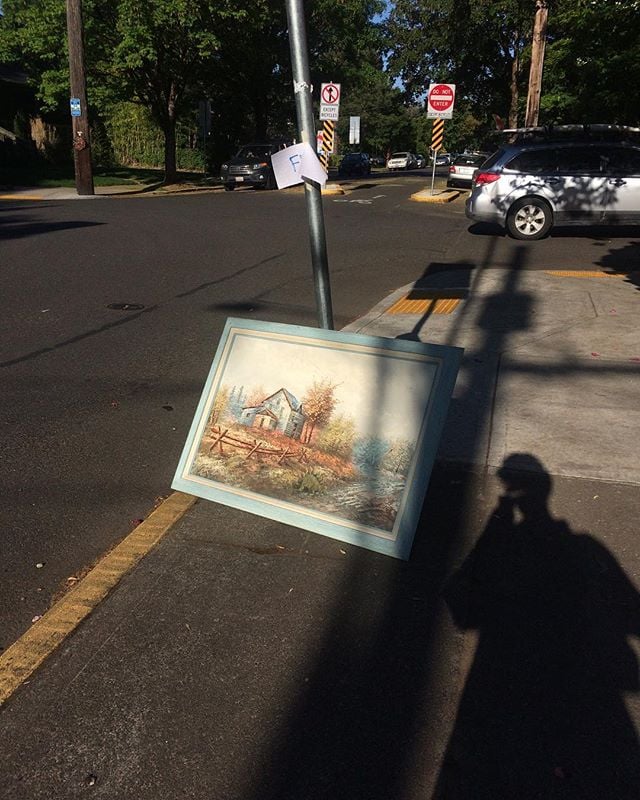 A large painting leans against a pole that has a "free" sign taped to it. The shadow of the photographer stretches down the sidewalk at it.