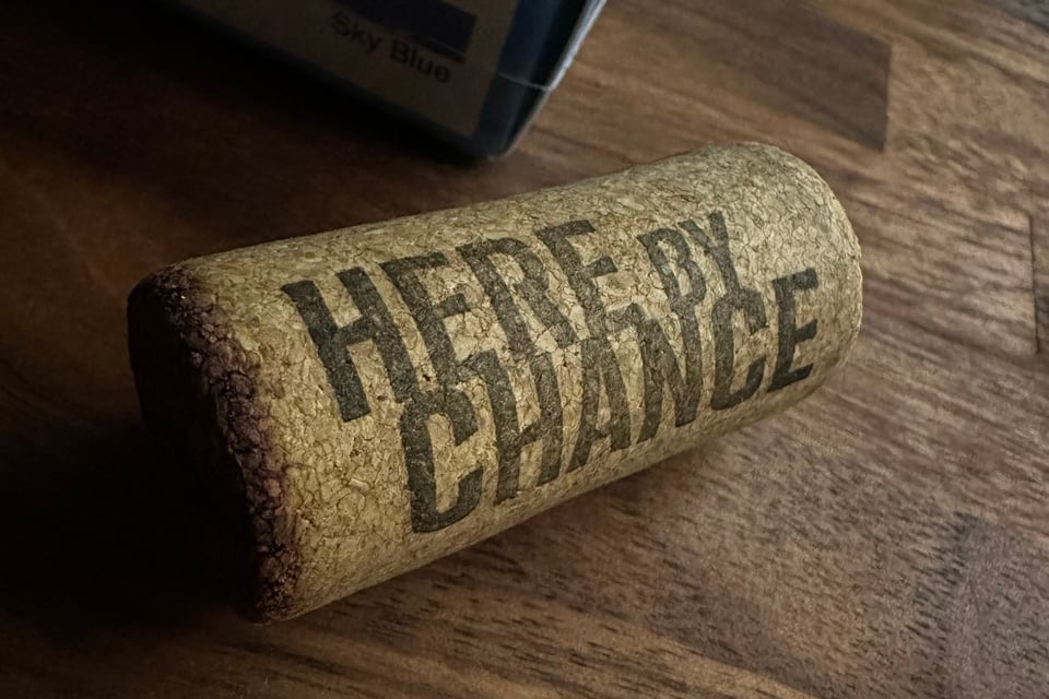 photo of wine-bottle cork on a butcher-block table; the cork has the words HERE BY CHANCE printed on it