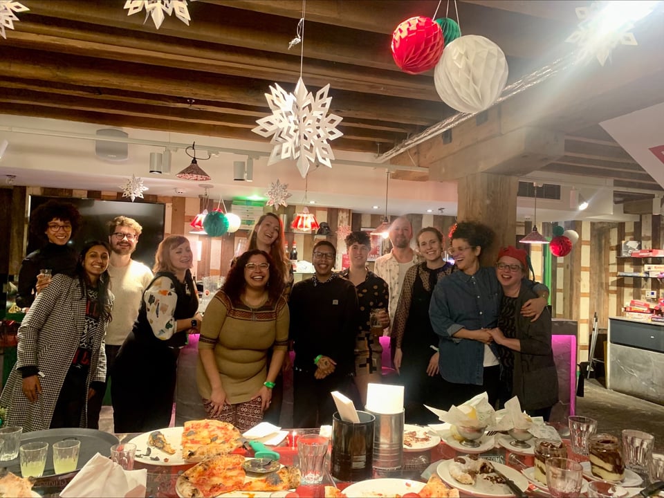 A group of 12 people standing in front of a table filled with food. The picture is taken in the middle of a Christmas party and everyone has stopped to have their photo taken. Lots of people are laughing.