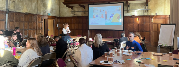 In a large room, we see a mixture of people sitting around tables, with coffee cups and paper plates. At the front of the room, a woman is presenting. A projector and pull down screen behind her shows an illustration of people working together.