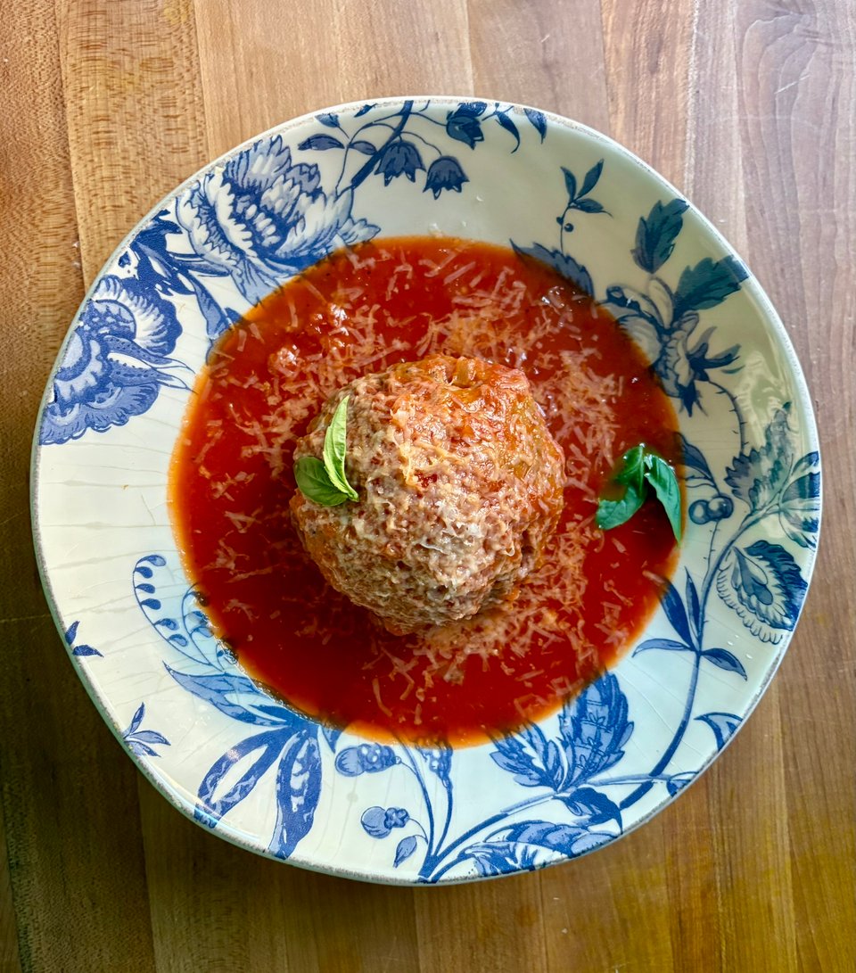Blue and white floral bowl containing a giant meatball in a tomato sauce topped with parmesan and basil leaves.