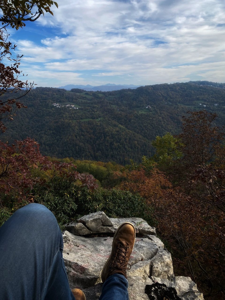 My feet and the rock I'm sitting on. On the background, an autumnal view of Slovenia with trees that are changing colors and mountains in the distance,