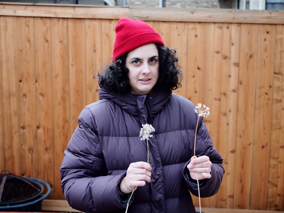 Portrait of Kat in a red beanie and purple coat, holding two stalks of dead plants