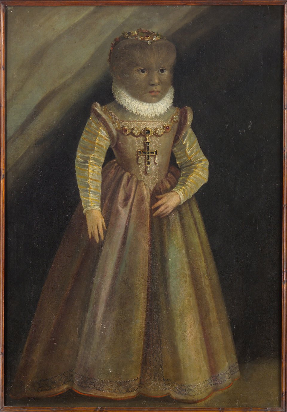 A little girl with a hairy face, wearing a shimmering pink dress that reaches her heels, stands against the mouth of a cave, looking serious. Around her neck is a thick white ruff and an enormous jewelled crucifix hands from the front of her dress.