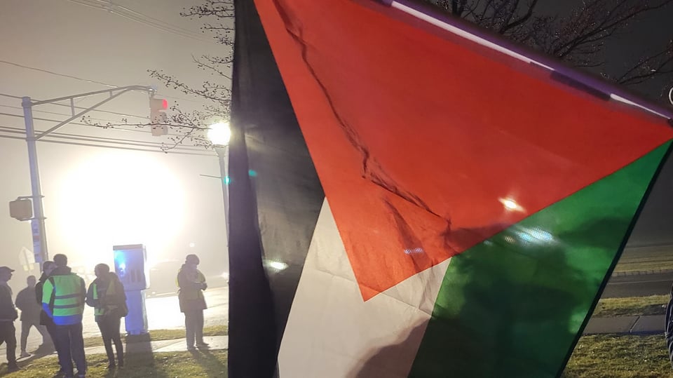 A Palestinian flag, backlit at night by bright lights is on the right side of the photo. On the left the misty air and bright lighting create a large white glow, with a traffic light and people standing in the middle ground between the light and the flag.