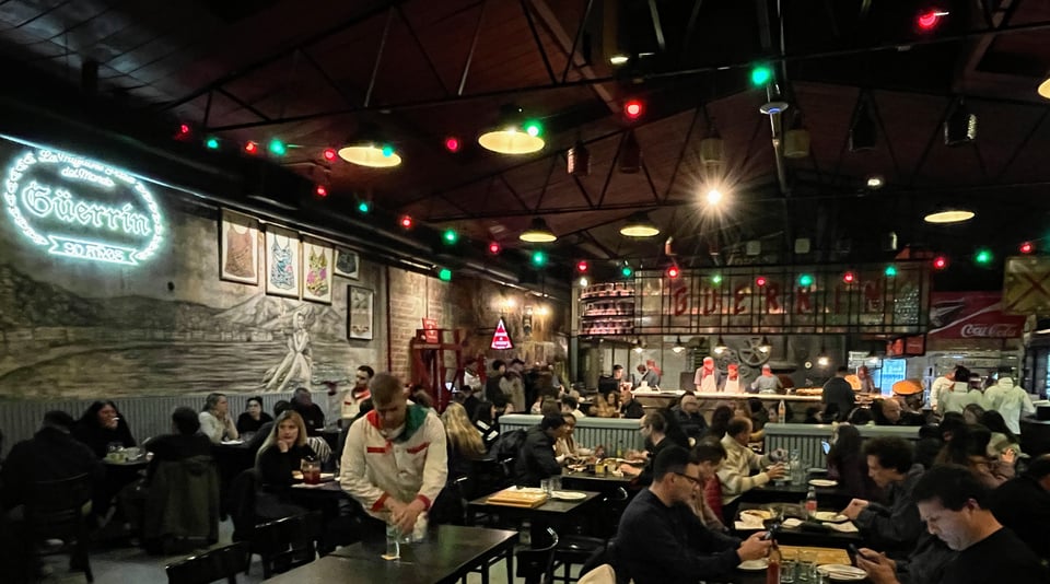 A bustling restaurant 'El Güerrín' with full tables, a neon sign of the restaurant on the wall and the pizza oven and cooks in the background.
