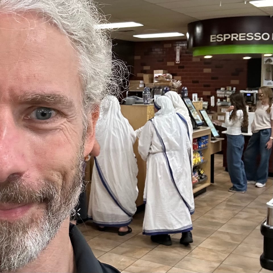 Selfie of half a face of a white man in a convenience store, with 3 white-robed nuns behind him