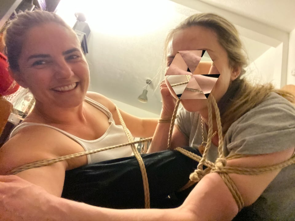 Two women in a room with ropes playfully tied around their bodies, connecting them together; one of the faces (Lesichka) is blurred and the other (Ariana) is smiling toward the camera.