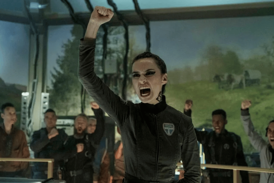 A screengrab from The Expanse, season 3. A pale skinned woman in heavy eyeliner and a jumpsuit raises a fist in a triumphant yell. She is surrounded by men in jumpsuits with their fists up, also yelling. This is Camina Drummer, played by Indigenous Canadian actor Cara Gee. She rules.