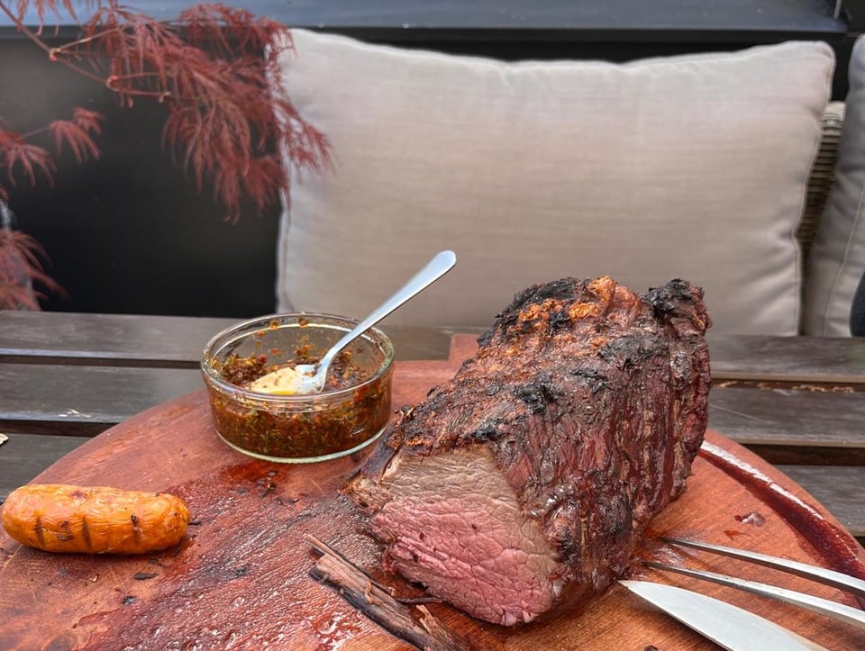 A hunk of roast beef and some sausages are on a wooden serving platter with a carving set laid next to them.