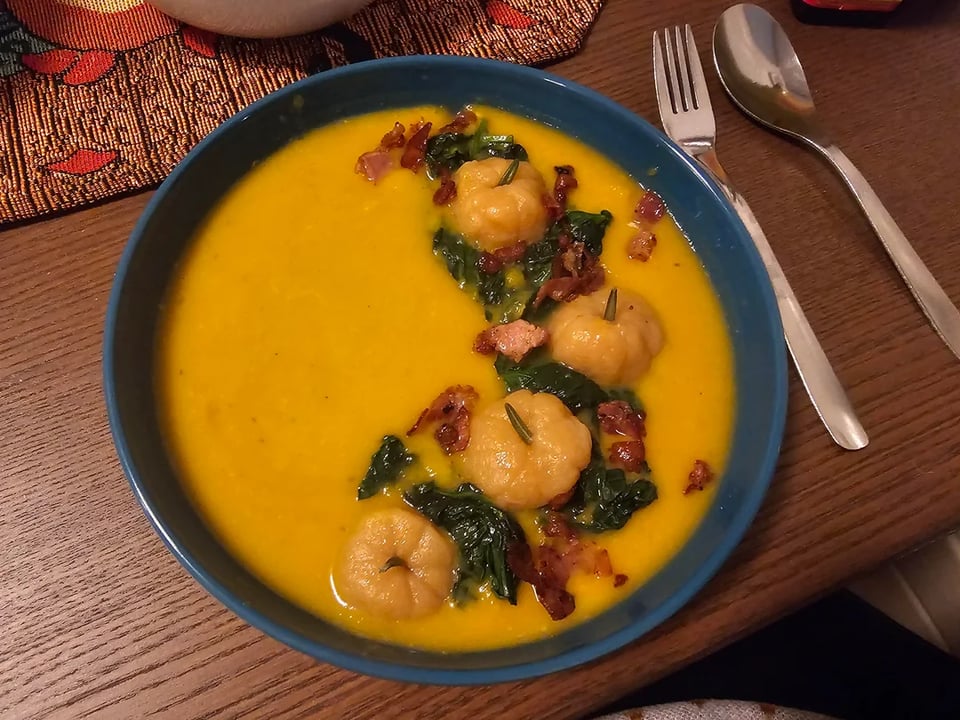 A bowl of pumpkin soup and a fork and spoon. There are pumpkin-like shapes in the soup, along with garnishes.