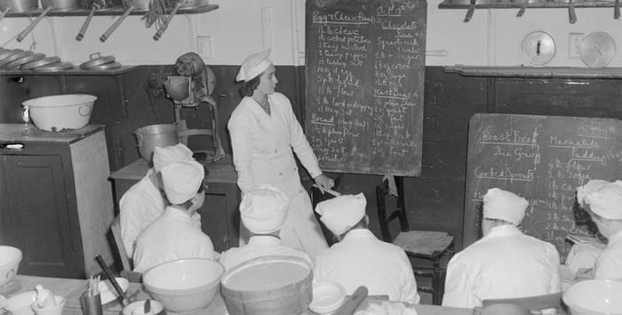 Trainee cooks during World War Two sit in front of a teacher who is going through the recipes for the day