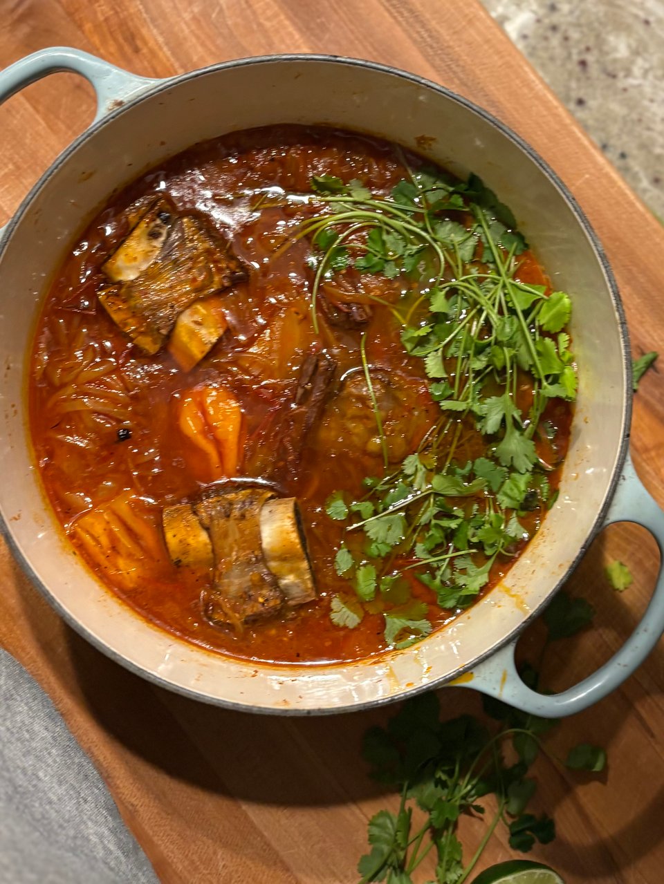 Big pot of clay-colored stew with beef short ribs, yucca, a habanero, and cilantro peeking out.