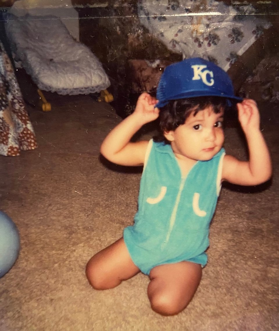 a slightly older Kandace in a blue jumper and playing with a royals baseball cap on her head