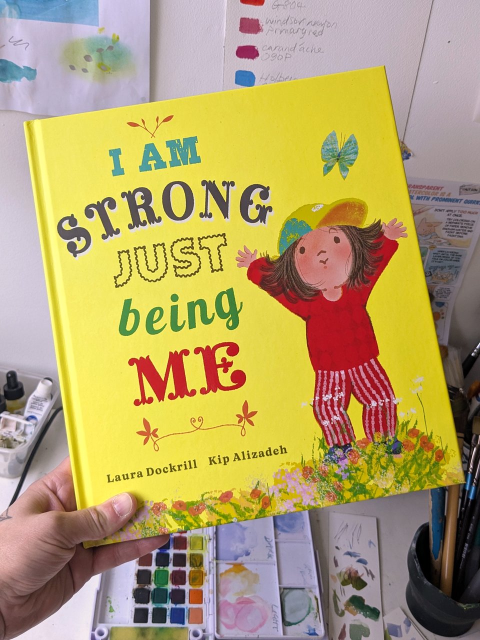A photo of the bright yellow front cover of I Am Strong Just Being Me a picture book by Laura Dockrill and Kip Alizadeh