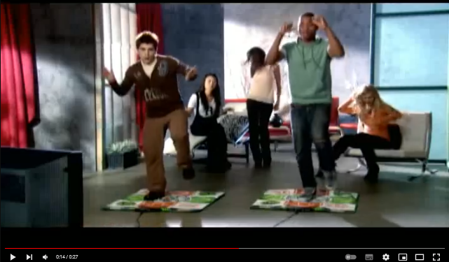 A screenshot from a trailer video for Dance Dance Revolution. Two young men dance on parallel dance pads. Three young women dance behind them, variously standing or seated. The image is as grainy as you might expect for the era.