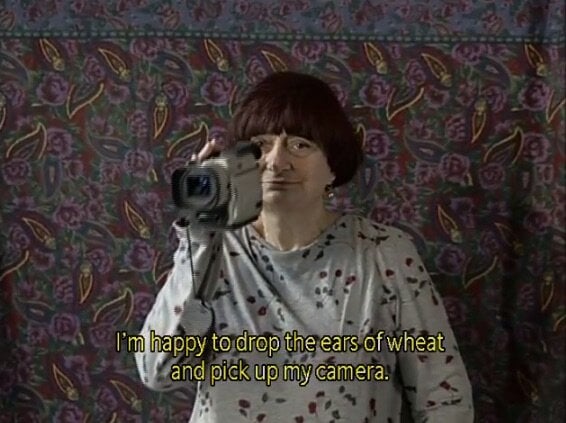 A film still of Agnés Varda holding a camera. She is standing in front of a paisley patterned backdrop. A caption beneath her says "I'm happy to drop the ears of what and pick up my camera."