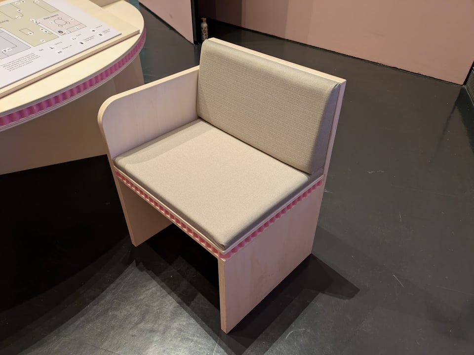 A wide chair with only one armrest sits next to a table. On both the table and chair, a small rim of soft pink foam wraps around the edge.