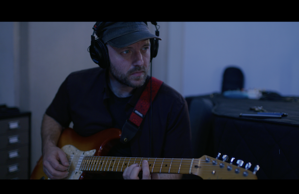 Carter Arrington, with guitar, looking pensive in the distance amidst a cool-lit backdrop