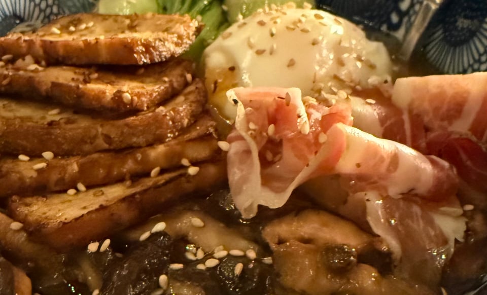 Close up on a ramen: shiitake, smoked tofu, soft egg, pork slices, carrots, paksoi and sesame seeds.