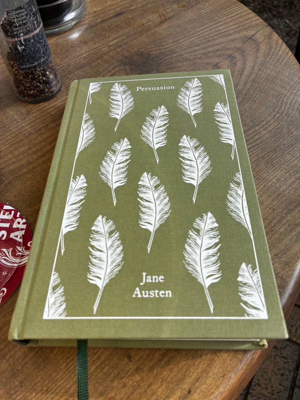 Image shows a cover of a book with a pattern of feathers, placed on a wooden table. The cover is olive in colour, the pattern is white. The book title and author are Persuasion and Jane Austen.