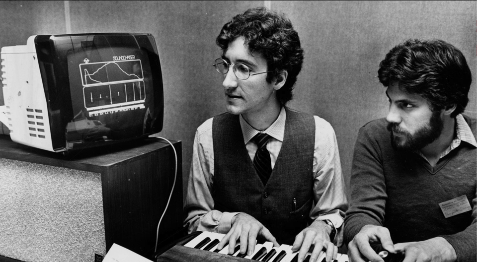 Two men at a keyboard stare at a computer monitor that is displaying musical notes and graphs.