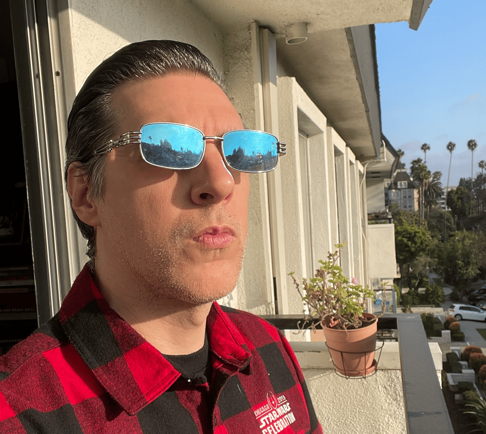 A man with blue reflective sunglasses stands on a balcony staring into the sun, buildings reflected in his glasses