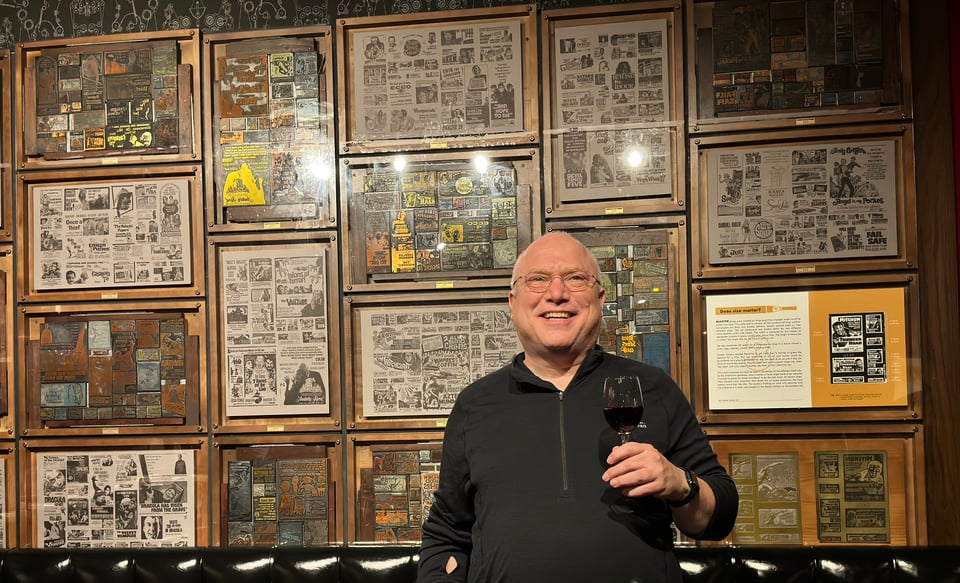 Photo of Glenn holding a glass of wine and smiling in front of a wall of framed small advertising printing plates and proof posters made from them