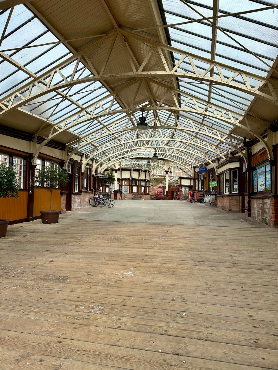 A Victoria railway station with broad cream painted roof supports and a wooden planked floor. There are some trees in large pots and some bikes in the distance. Image by Rowan Ambrose.