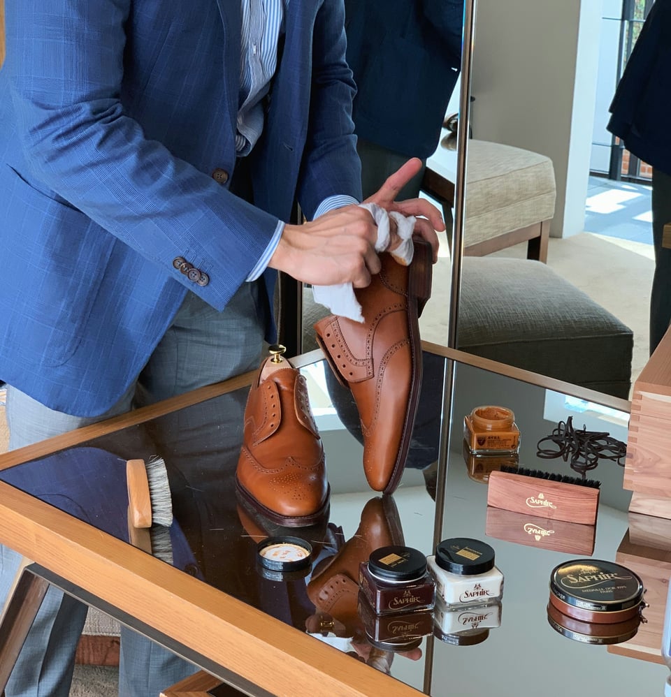 A white person in a blue suit cleans brown leather shoes. Leather cleaning supplies are also visible