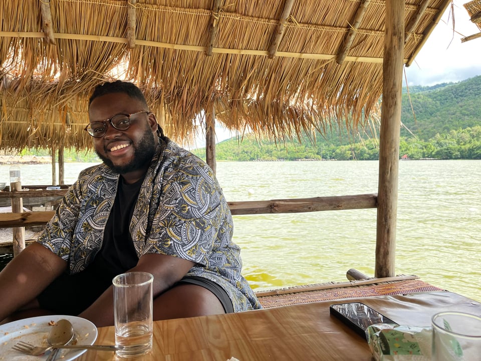 a black man smiles, sitting in a thatched roof hut, water and trees stretch out behind him
