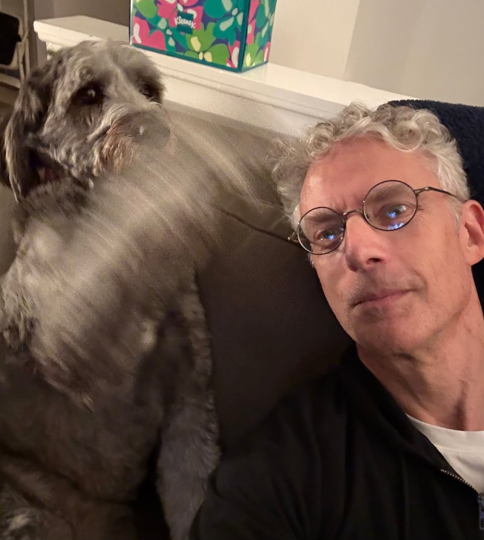 Selfie of middle-aged white man with round glasses and gray aussiedoodle on a sofa; the dog's from left leg is in motion and an instant away from striking the man in the face