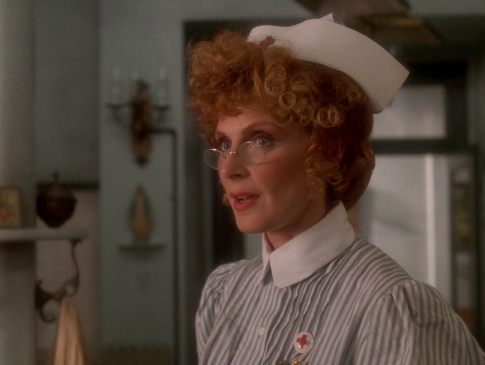 Doctor Crusher dressed in a 19th century nurses uniform. Her hair is up and she's wearing glasses.