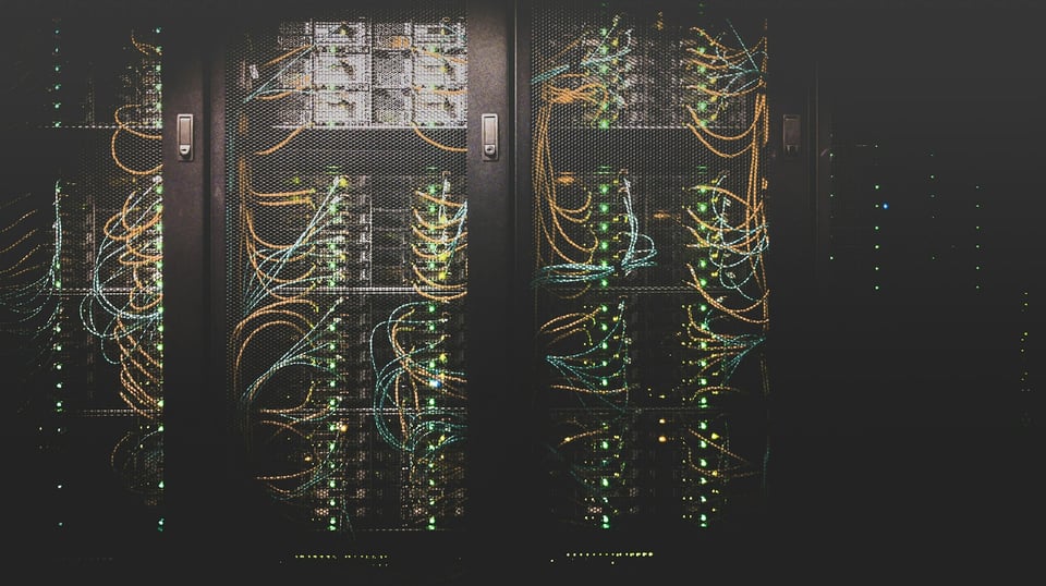 A wall of racked machines in a server room cage. Three cages are well-lit; the fourth is dim. Photo by Taylor Vick on Unsplash