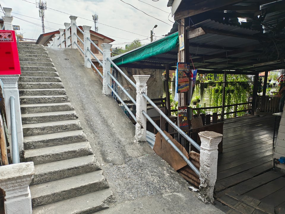 A bridge over a canal in the Artist's House in Bangkok, a large traditional house now converted into galleries and cafes. On the left side of the image, a concrete bridge with steps and a ramp lead over the canal, filled with brownish green water. Turn toward the right and the path becomes a covered wooden terrace overlooking the canal, with a wooden railing and plants hanging from above.