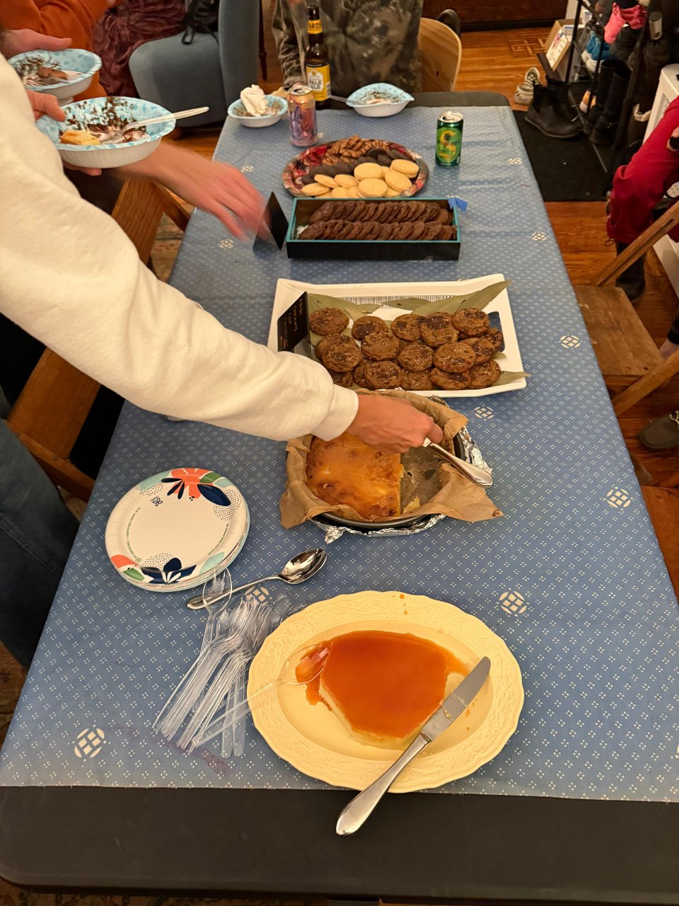 a table with a blue tablecloth and people reaching for dishes on the table, all desserts. cookies, leche flan, cassava cake