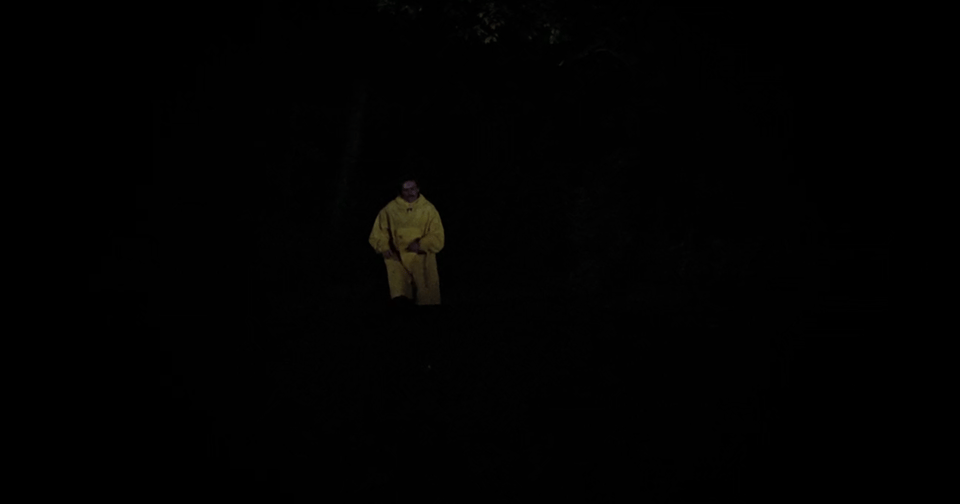 Steve runs towards the camera in the dark, the hood of his yellow rain coat down. At the very top of the screen, one can just barely make out leaves hanging down from a tree. Everything else is black.