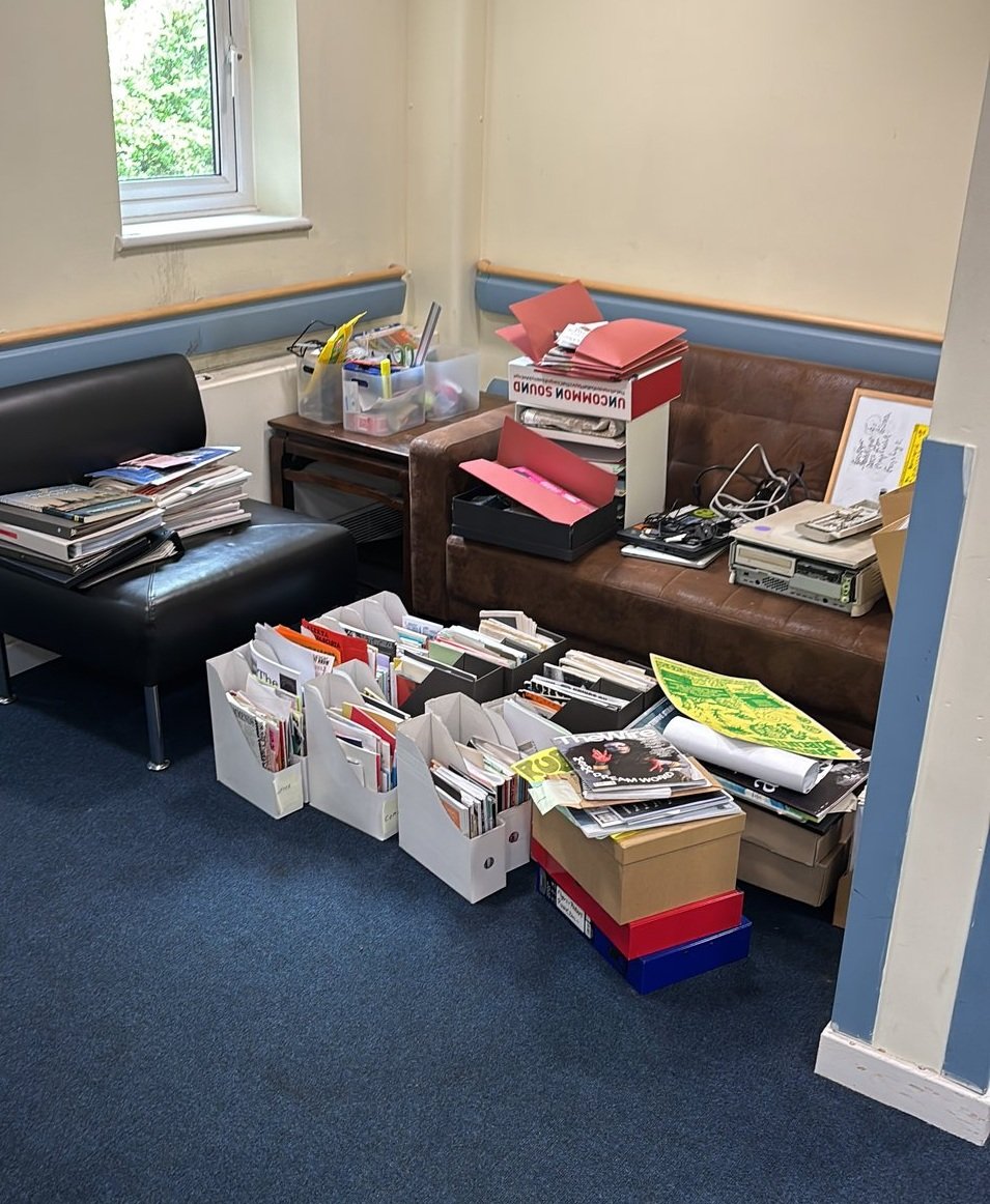 A cluttered workspace featuring a black sofa, a variety of stacked papers, and several boxes filled with magazines, books, and supplies.