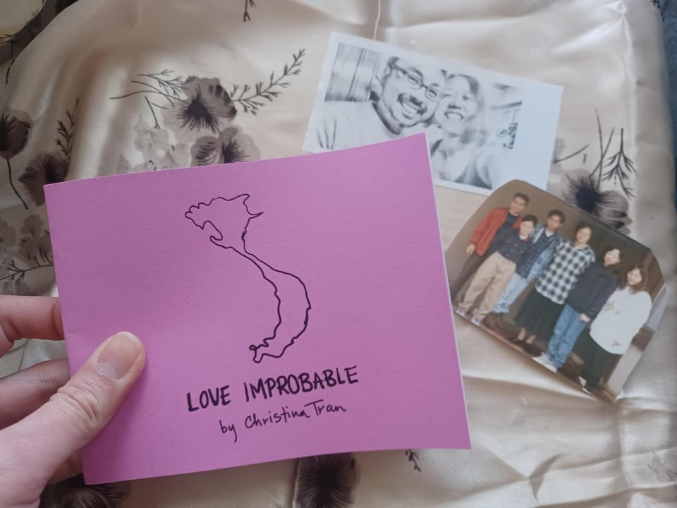 A hand holds purple quarter-sized zine entitled "Love Improbable" featuring an illustrated silhouette of the country of Vietnam. In the background are family photos against a tan silk scarf.