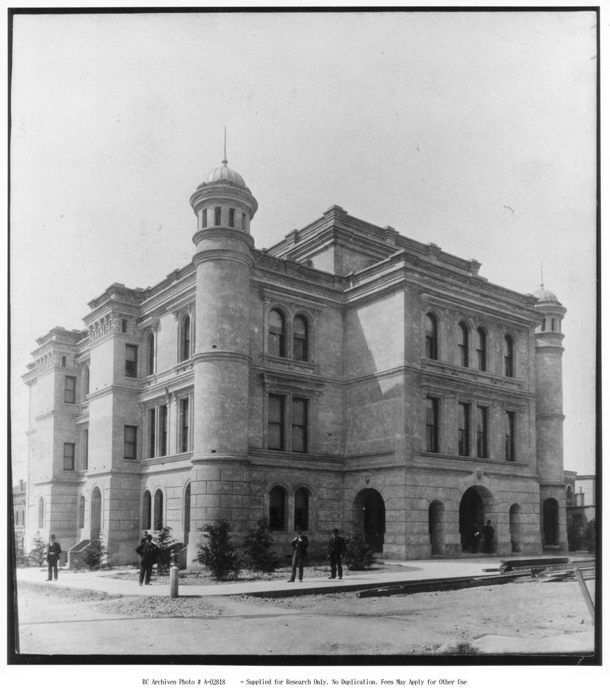 Black and white photo. A group of men stand ins front of a large building, featuring a arches and turrets.