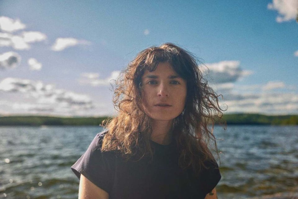 A portrait of a woman looking into the camera. She stands in front of a body of water, and her curly shoulder-length brown hair looks windswept.