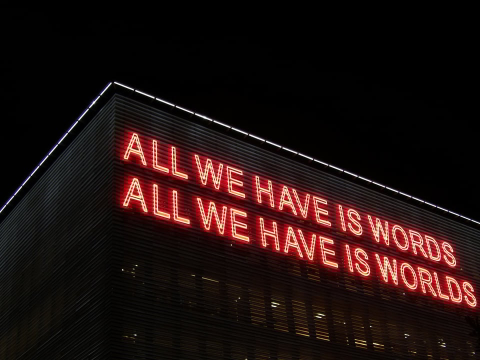 Red neon sign lit on a building at night reads: "All we have is words. All we have is worlds" The building has white lights along the roof line, barely outlining the dark building against the black sky.