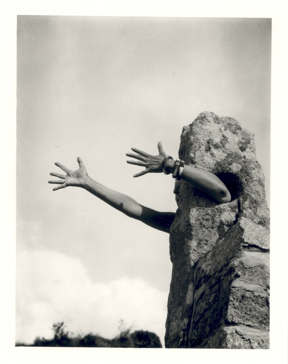 A black-and-white vertical photo shows an outstretched pair of arms emerging from a rough, human-scale obelisk abutting a stone-and-mortar column or wall on the right side of the composition. In the distance, scruffy plantlife at the horizon, and a pale sky of low, puffy clouds.