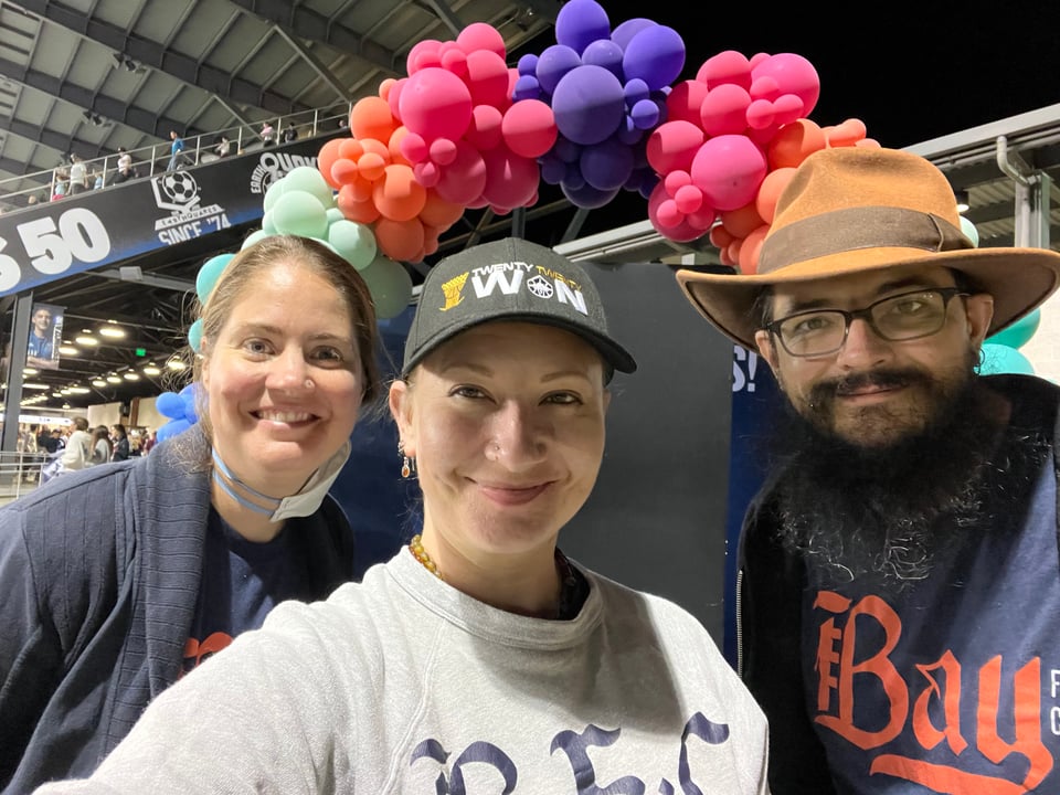 three people smile at the camera from in front of a rainbow balloon arch. they are all three wearing Bay FC shirts and look like friends.