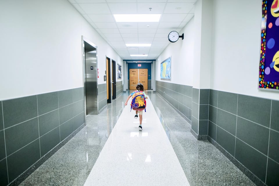 Little girl walking down a long school hallway, photo is from the back